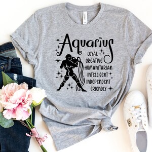 May include: A heather gray t-shirt featuring a black graphic of the Aquarius zodiac sign, a water bearer figure, and the words Aquarius, Loyal, Creative, Humanitarian, Intelligent, Independent, and Friendly, surrounded by small star icons.