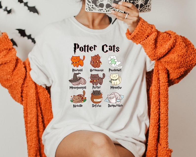 May include: White t-shirt with a graphic of cats representing characters from the Harry Potter series. The text "Potter Cats" is at the top of the graphic. The names of the characters are listed below each cat.
