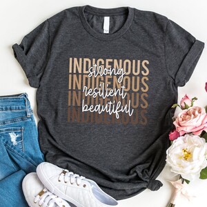 Może przedstawiać: Szary t-shirt z nadrukiem "Indigenous strong resilient beautiful" w kolorze białym na przodzie. Słowa są ułożone jedno nad drugim i są w gradiencie brązowych odcieni.