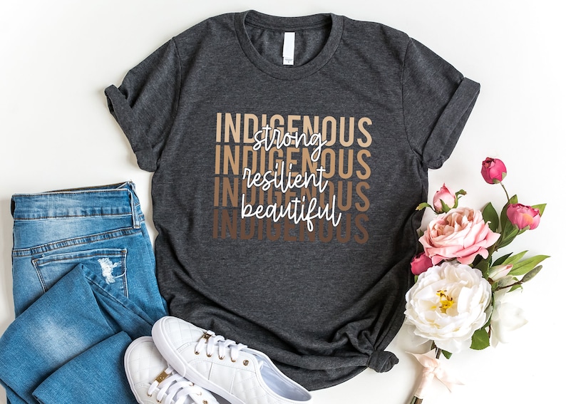 Może przedstawiać: Szary t-shirt z nadrukiem "Indigenous strong resilient beautiful" w kolorze białym na przodzie. Słowa są ułożone jedno nad drugim i są w gradiencie brązowych odcieni.