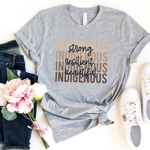 Może przedstawiać: Szary T-shirt z napisami "Indigenous", "strong", "resilient", "beautiful" i "Indigenous" ułożonymi jeden nad drugim w brązowych i beżowych literach.