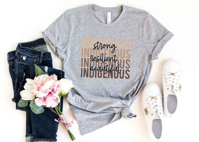 Może przedstawiać: Szary T-shirt z napisami "Indigenous", "strong", "resilient", "beautiful" i "Indigenous" ułożonymi jeden nad drugim w brązowych i beżowych literach.