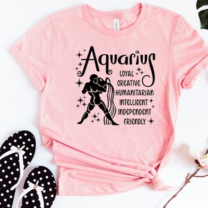 May include: A light pink t-shirt featuring a black graphic of the Aquarius zodiac sign, a figure pouring water, and the words Aquarius, Loyal, Creative, Humanitarian, Intelligent, Independent, and Friendly, accented with small star symbols.