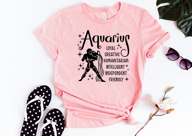 May include: A light pink t-shirt featuring a black graphic of the Aquarius zodiac sign, a figure pouring water, and the words Aquarius, Loyal, Creative, Humanitarian, Intelligent, Independent, and Friendly, accented with small star symbols.