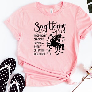 Op de afbeelding: Roze T-shirt met een zwarte afbeelding van een Boogschutter sterrenbeeld en de woorden "Boogschutter", "Onafhankelijk", "Grootmoedig", "Zorgzaam", "Eerlijk", "Optimistisch" en "Intelligent".