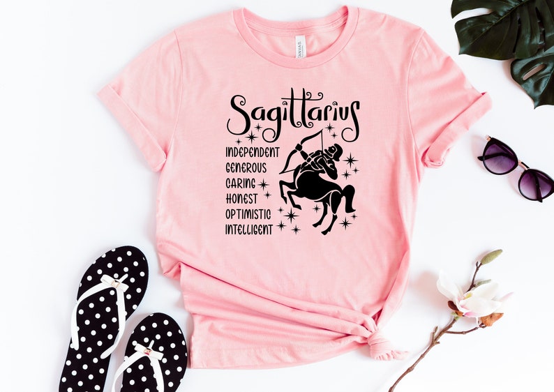 Op de afbeelding: Roze T-shirt met een zwarte afbeelding van een Boogschutter sterrenbeeld en de woorden "Boogschutter", "Onafhankelijk", "Grootmoedig", "Zorgzaam", "Eerlijk", "Optimistisch" en "Intelligent".