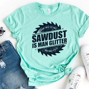 Pu&ograve; includere: Una maglietta blu chiaro con una grafica a forma di lama da sega che dice "MEASURE TWICE CUT ONCE SAWDUST IS MAN GLITTER THEN FORCE IT TO FIT" in testo nero.