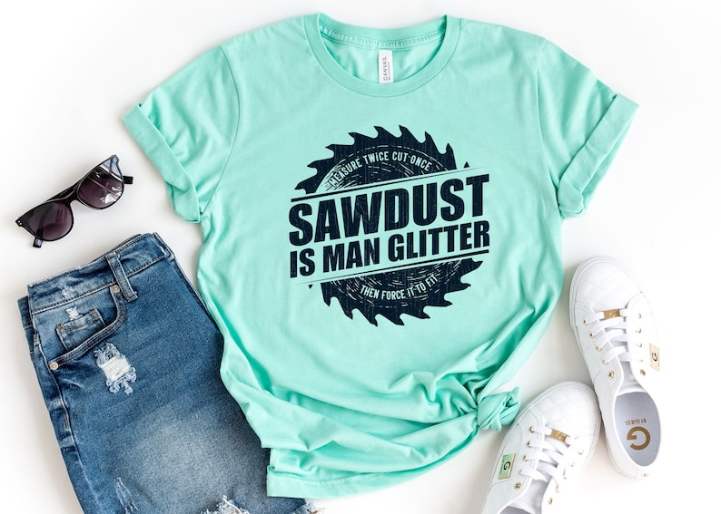 Pu&ograve; includere: Una maglietta blu chiaro con una grafica a forma di lama da sega che dice "MEASURE TWICE CUT ONCE SAWDUST IS MAN GLITTER THEN FORCE IT TO FIT" in testo nero.