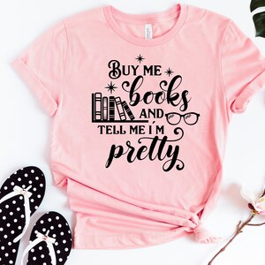 Puede incluir: Camiseta rosa con texto negro que dice "C&oacute;mprame libros y dime que soy bonita". El texto est&aacute; rodeado de estrellas e incluye una ilustraci&oacute;n de una estanter&iacute;a con libros y gafas.