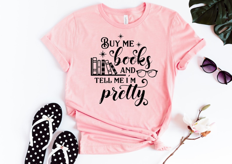 Puede incluir: Camiseta rosa con texto negro que dice "C&oacute;mprame libros y dime que soy bonita". El texto est&aacute; rodeado de estrellas e incluye una ilustraci&oacute;n de una estanter&iacute;a con libros y gafas.