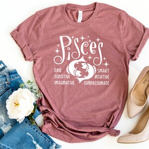 Op de afbeelding: Een mauvekleurig T-shirt met het woord "Pisces" in witte letters, met een visafbeelding. Het shirt vermeldt eigenschappen: vriendelijk, gevoelig, fantasierijk, slim, intu&iuml;tief en meelevend. Het shirt wordt gecombineerd met een blauwe jeans en beige hoge hakken.