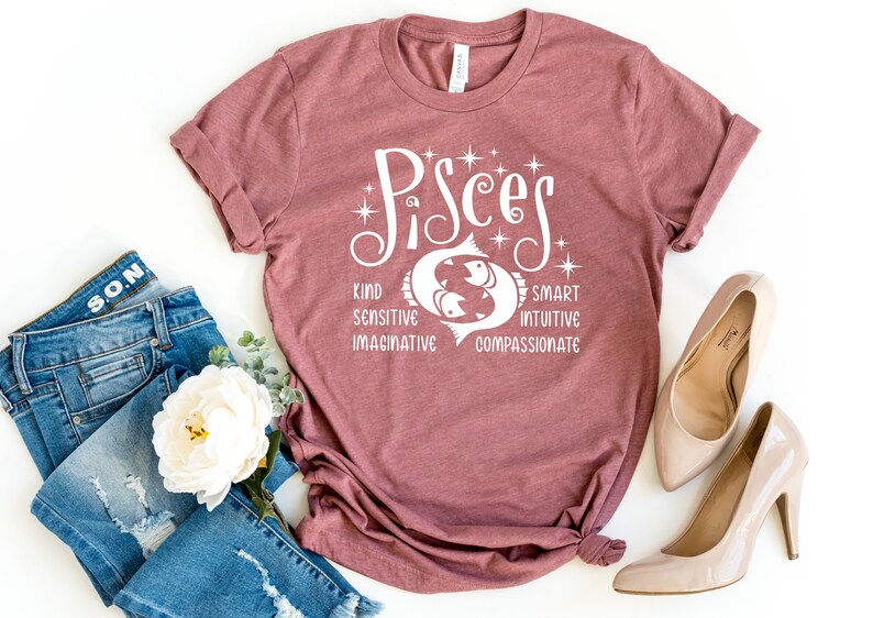 Op de afbeelding: Een mauvekleurig T-shirt met het woord "Pisces" in witte letters, met een visafbeelding. Het shirt vermeldt eigenschappen: vriendelijk, gevoelig, fantasierijk, slim, intu&iuml;tief en meelevend. Het shirt wordt gecombineerd met een blauwe jeans en beige hoge hakken.