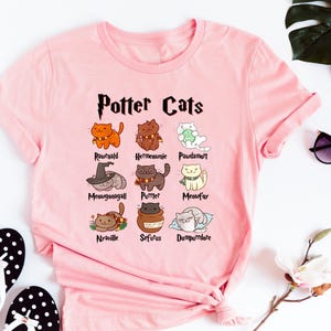 May include: Pink t-shirt with a graphic of cats representing characters from the Harry Potter series. The text "Potter Cats" is at the top of the graphic. The names of the characters are listed below each cat.