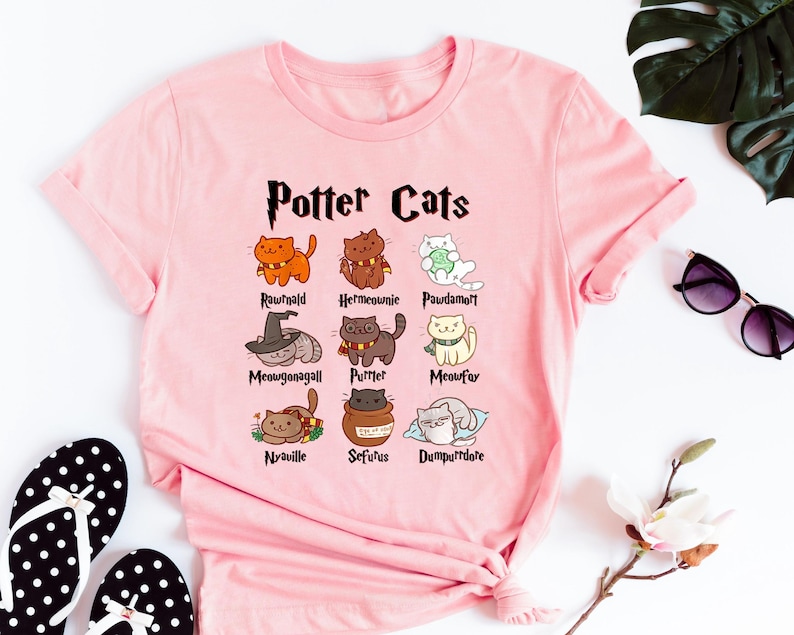 May include: Pink t-shirt with a graphic of cats representing characters from the Harry Potter series. The text "Potter Cats" is at the top of the graphic. The names of the characters are listed below each cat.