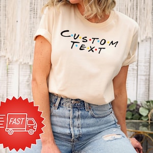 May include: A cream-coloured t-shirt featuring the text "CUSTOM TEXT" in black with colourful dots. The t-shirt is worn with blue jeans. A red sticker with a white truck and the word "FAST" is in the lower left corner.