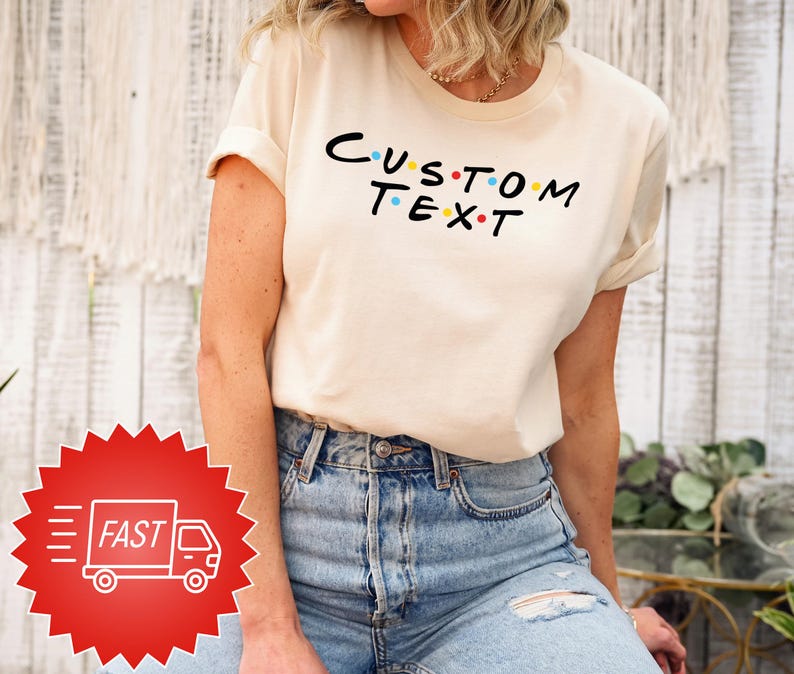 May include: A cream-coloured t-shirt featuring the text "CUSTOM TEXT" in black with colourful dots. The t-shirt is worn with blue jeans. A red sticker with a white truck and the word "FAST" is in the lower left corner.