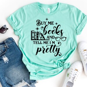 Puede incluir: Una camiseta turquesa con el texto "Buy me books and tell me I'm pretty" en negro. El texto est&aacute; rodeado por una pila de libros, gafas y estrellas.