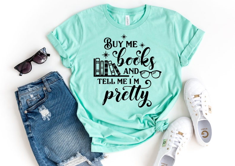 Puede incluir: Una camiseta turquesa con el texto "Buy me books and tell me I'm pretty" en negro. El texto est&aacute; rodeado por una pila de libros, gafas y estrellas.