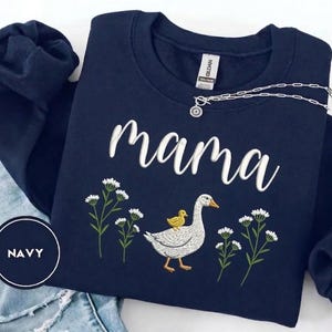 May include: Navy blue sweatshirt with the word "mama" in white script. The design includes a goose with a yellow chick on its back, surrounded by white flowers. A silver necklace is draped across the sweatshirt.