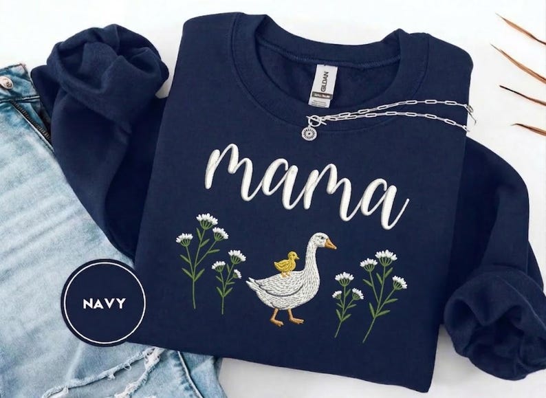 May include: Navy blue sweatshirt with the word "mama" in white script. The design includes a goose with a yellow chick on its back, surrounded by white flowers. A silver necklace is draped across the sweatshirt.