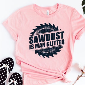 Pu&ograve; includere: Maglietta rosa con una grafica di lama da sega nera che dice "Sawdust is Man Glitter" e "Measure Twice Cut Once Then Force It To Fit".