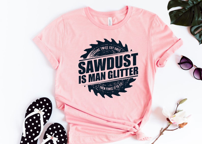 Pu&ograve; includere: Maglietta rosa con una grafica di lama da sega nera che dice "Sawdust is Man Glitter" e "Measure Twice Cut Once Then Force It To Fit".