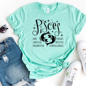 Op de afbeelding: Een lichtblauwgroen t-shirt met het woord "Pisces" in zwart schrift, met een Vissen-symbool en bijbehorende eigenschappen. Het shirt is gestyled met een blauwe spijkerbroek, zonnebril en witte sneakers.