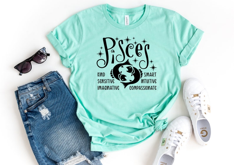 Op de afbeelding: Een lichtblauwgroen t-shirt met het woord "Pisces" in zwart schrift, met een Vissen-symbool en bijbehorende eigenschappen. Het shirt is gestyled met een blauwe spijkerbroek, zonnebril en witte sneakers.