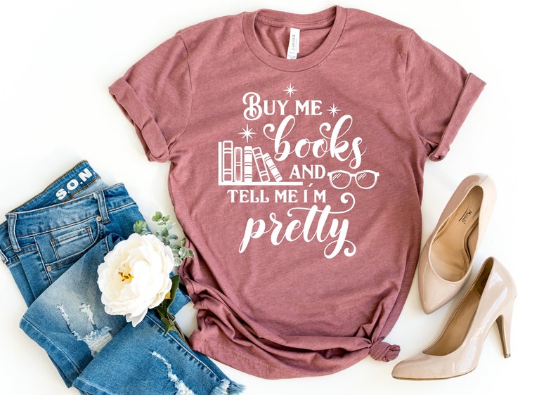 Puede incluir: Una camiseta de color marr&oacute;n rojizo con el texto "Buy me books and tell me I'm pretty" en blanco. El texto est&aacute; rodeado de estrellas e incluye una pila de libros y un par de gafas.