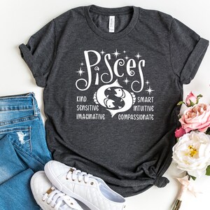 Op de afbeelding: Donkergrijs t-shirt met het woord "Pisces" in witte letters, met een vissymbool en de woorden "Kind, Sensitive, Imaginative, Smart, Intuitive, Compassionate." Het shirt wordt gecombineerd met een blauwe jeans en witte sneakers. Er is ook een boeket roze en witte rozen aanwezig.
