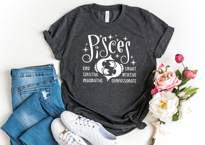 Op de afbeelding: Donkergrijs t-shirt met het woord "Pisces" in witte letters, met een vissymbool en de woorden "Kind, Sensitive, Imaginative, Smart, Intuitive, Compassionate." Het shirt wordt gecombineerd met een blauwe jeans en witte sneakers. Er is ook een boeket roze en witte rozen aanwezig.