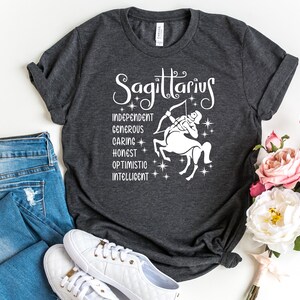 Op de afbeelding: Een donkergrijs T-shirt met een wit grafisch ontwerp met het sterrenbeeld Boogschutter en de woorden "Boogschutter", "Onafhankelijk", "Grootmoedig", "Zorgzaam", "Eerlijk", "Optimistisch" en "Intelligent".
