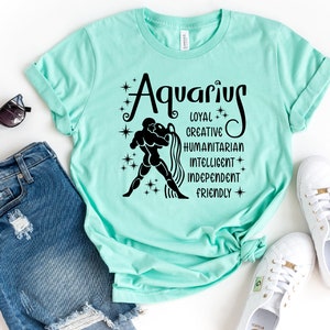 May include: A mint green t-shirt featuring a black graphic of the Aquarius zodiac sign, a figure pouring water, and the words Aquarius, Loyal, Creative, Humanitarian, Intelligent, Independent, and Friendly, surrounded by small star icons.