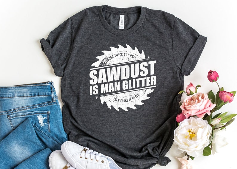 Pu&ograve; includere: Una maglietta grigio scuro con una grafica bianca di una lama da sega che dice "Measure Twice Cut Once Sawdust Is Man Glitter Then Force It To Fit".