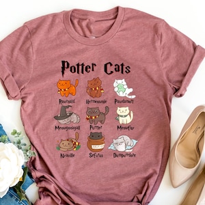 May include: A maroon t-shirt with the text "Potter Cats" printed on the front. The shirt features a series of cartoon cats with names like "Rougald", "Hermione", and "Paurdamott".