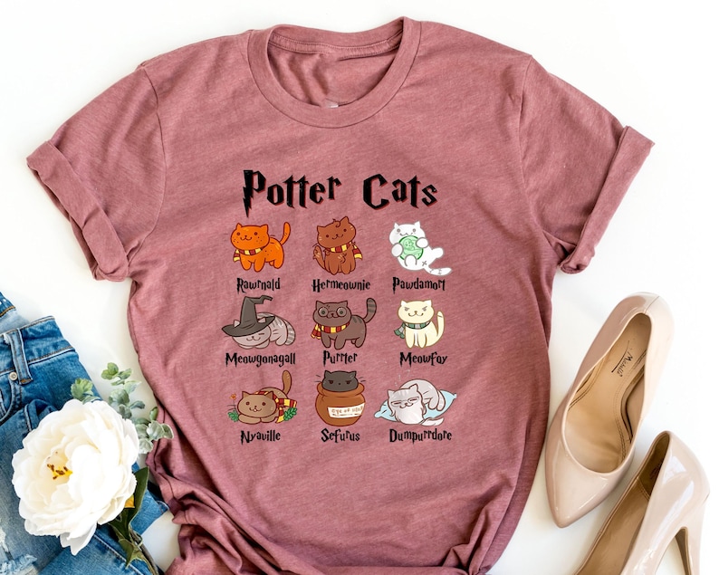 May include: A maroon t-shirt with the text "Potter Cats" printed on the front. The shirt features a series of cartoon cats with names like "Rougald", "Hermione", and "Paurdamott".
