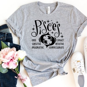 Op de afbeelding: Een lichtgrijs t-shirt met het woord "Pisces" in zwart schrift, met een Vissen-symbool en de woorden "Vriendelijk, Gevoelig, Fantasierijk, Slim, Intu&iuml;tief, Medelevend". Het shirt wordt gecombineerd met een blauwe jeans, witte sneakers en een boeket roze en witte bloemen.