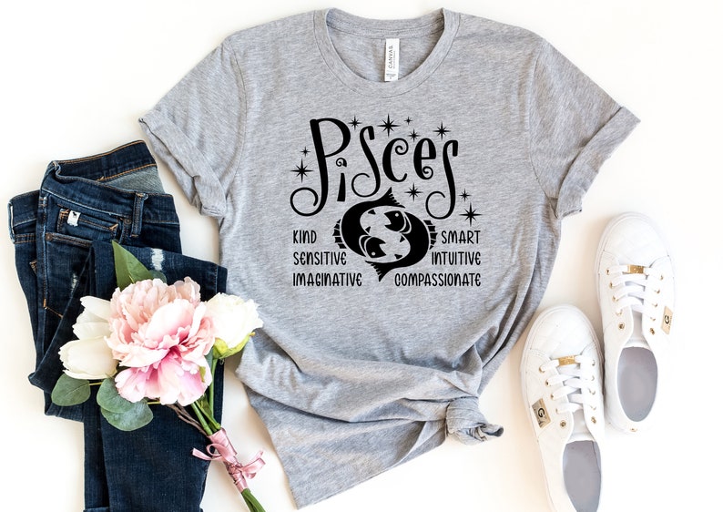 Op de afbeelding: Een lichtgrijs t-shirt met het woord "Pisces" in zwart schrift, met een Vissen-symbool en de woorden "Vriendelijk, Gevoelig, Fantasierijk, Slim, Intu&iuml;tief, Medelevend". Het shirt wordt gecombineerd met een blauwe jeans, witte sneakers en een boeket roze en witte bloemen.