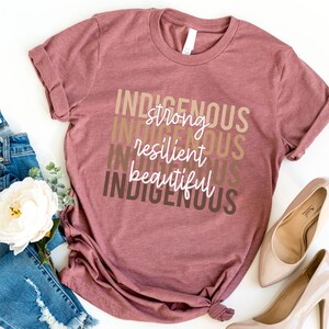 Może przedstawiać: Maronowy T-shirt z tekstem "INDIGENOUS" powt&oacute;rzonym cztery razy w r&oacute;żnych odcieniach brązu. Tekst jest ułożony pionowo, a między słowami "INDIGENOUS" znajdują się słowa "strong", "resilient" i "beautiful" w białym kolorze.