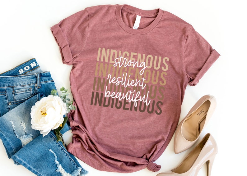 Może przedstawiać: Maronowy T-shirt z tekstem "INDIGENOUS" powt&oacute;rzonym cztery razy w r&oacute;żnych odcieniach brązu. Tekst jest ułożony pionowo, a między słowami "INDIGENOUS" znajdują się słowa "strong", "resilient" i "beautiful" w białym kolorze.