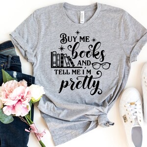 Puede incluir: Una camiseta gris con el texto "Buy me books and tell me I'm pretty" en letras negras. El texto est&aacute; rodeado de estrellas e incluye una pila de libros y un par de gafas.