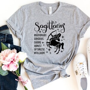 Op de afbeelding: Grijs T-shirt met een zwarte afbeelding van het sterrenbeeld Boogschutter en de woorden "Boogschutter", "Onafhankelijk", "Grootmoedig", "Zorgzaam", "Eerlijk", "Optimistisch" en "Intelligent".