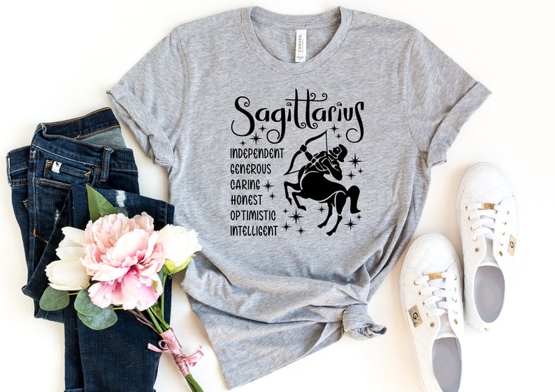 Op de afbeelding: Grijs T-shirt met een zwarte afbeelding van het sterrenbeeld Boogschutter en de woorden "Boogschutter", "Onafhankelijk", "Grootmoedig", "Zorgzaam", "Eerlijk", "Optimistisch" en "Intelligent".