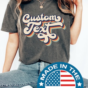 May include: Dark grey t-shirt featuring the words "Custom Text" in a retro font with a rainbow colour scheme. Made in the USA.
