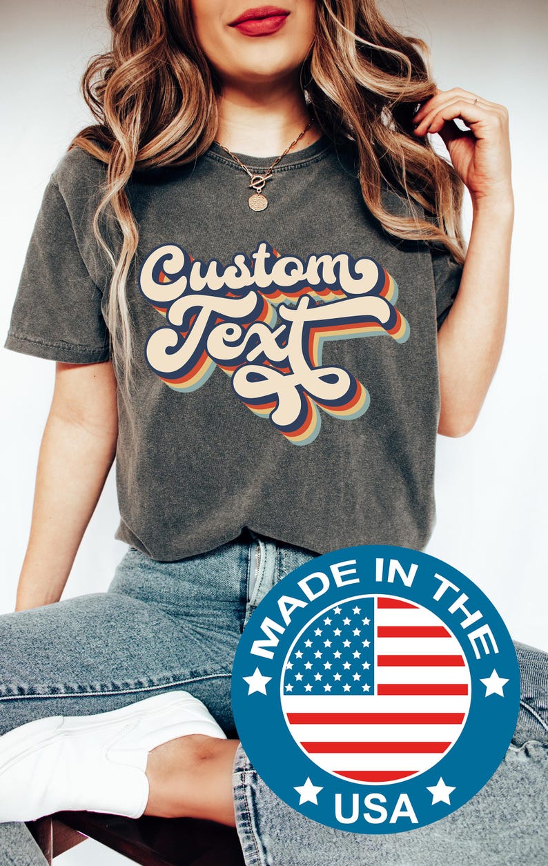 May include: Dark grey t-shirt featuring the words "Custom Text" in a retro font with a rainbow colour scheme. Made in the USA.