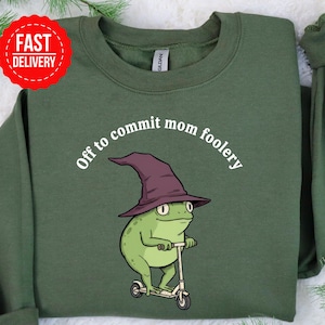 May include: Green sweatshirt with the phrase "Off to commit mom foolery" above a cartoon frog wearing a witch hat and riding a scooter. A red and white "Fast Delivery" sticker is in the upper left corner.