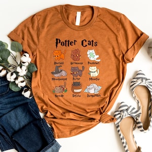 May include: A brown t-shirt with the text "Potter Cats" and illustrations of cats with names like "Ronweald", "Hermione", "Padma", "Neville", "Sirius", and "Dumbledore".