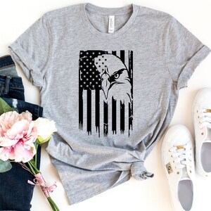 Może przedstawiać: Szary T-shirt z czarno-białym nadrukiem orła przed amerykańską flagą.