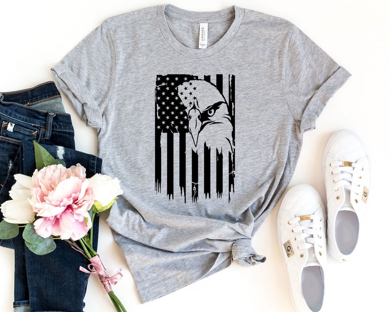 Może przedstawiać: Szary T-shirt z czarno-białym nadrukiem orła przed amerykańską flagą.