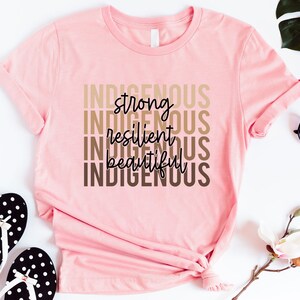 Może przedstawiać: R&oacute;żowa koszulka z nadrukiem "Indigenous strong resilient beautiful Indigenous" w kolorze brązowym i czarnym na przodzie.